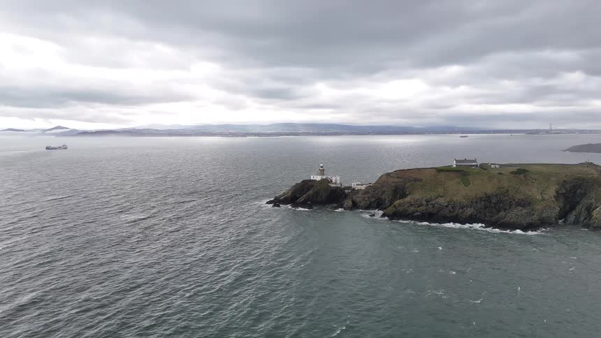 A still aerial view of a historic lighthouse perched on a rugged, grassy headland surrounded by the choppy sea under a dramatic, overcast sky