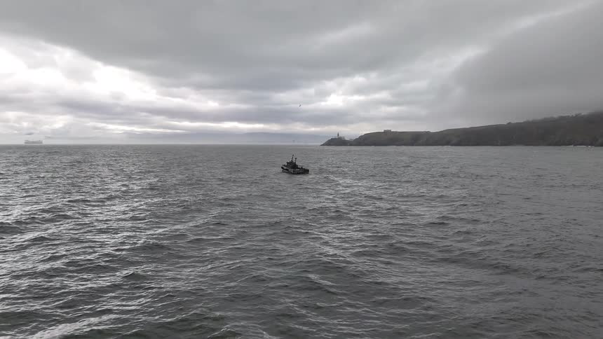 An aerial shot slowly pulls away from a lone boat on a vast, choppy grey sea, revealing a distant lighthouse on the misty coastline under an overcast sky