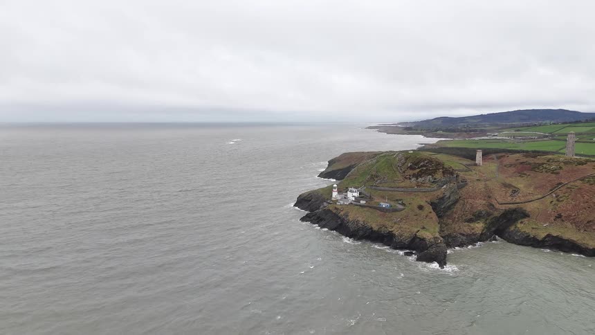A slow aerial pullback reveals the Wicklow Head Lighthouse and historic towers standing guard on the rugged Irish coast under a grey, overcast sky
