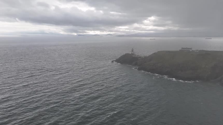 An atmospheric aerial view shows a lighthouse standing guard on a rocky headland as a storm brews over the gray sea