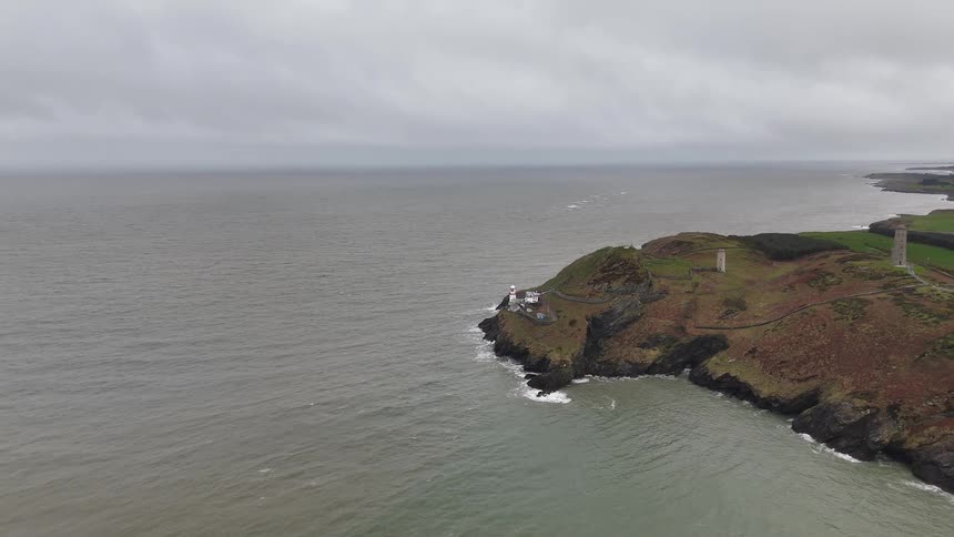 A slow aerial pullback reveals a lighthouse and stone towers on a rugged point overlooking a moody sea on an overcast day