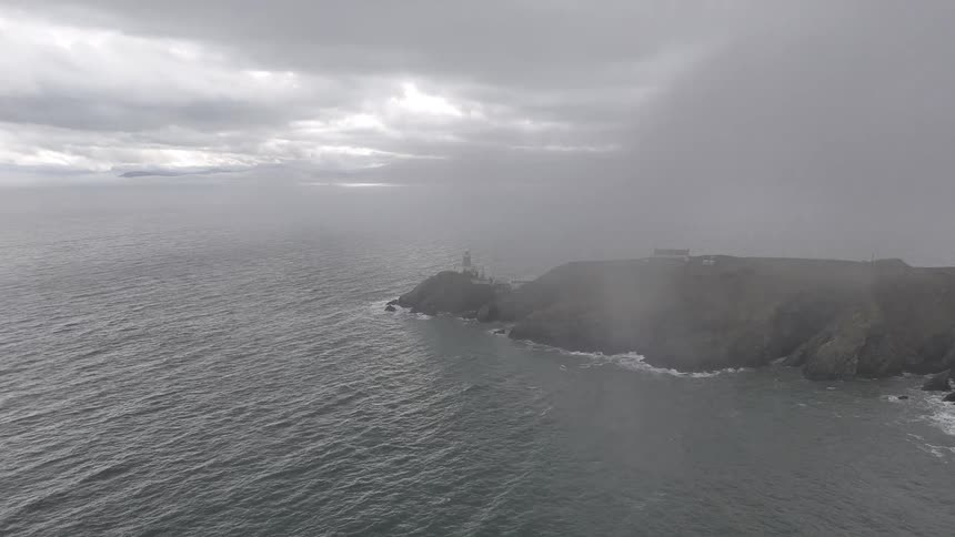 An atmospheric aerial shot flies through thick grey fog to reveal a lighthouse on a rugged cliffside over the ocean