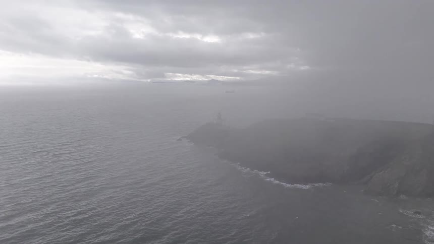An aerial shot pushes through thick fog to reveal a lighthouse on a rugged cliffside overlooking the sea
