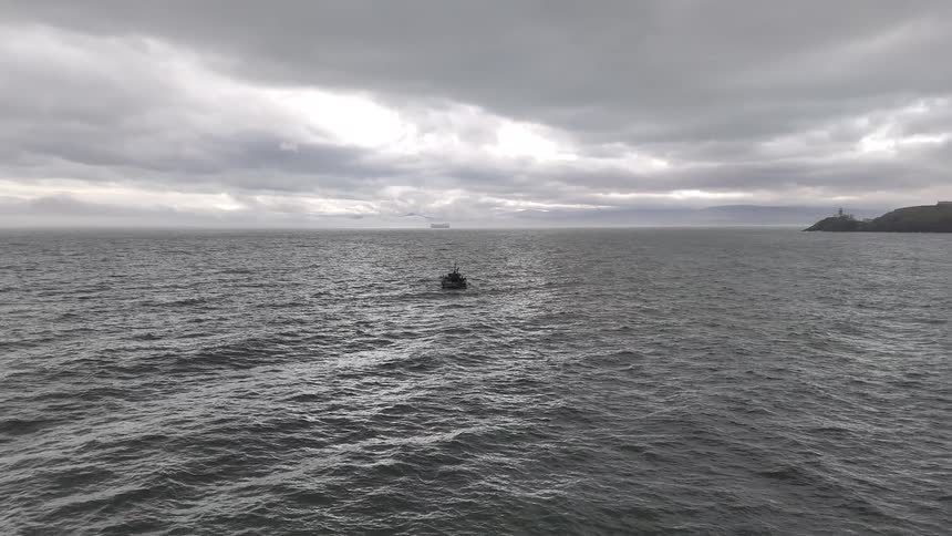 A solitary fishing trawler drifts on a vast, choppy grey sea under a heavy overcast sky, with a distant coastline visible on the horizon