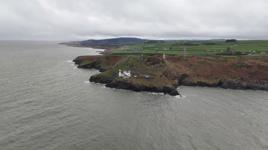 A wide, static aerial shot captures a lighthouse on a rugged headland overlooking the sea, with rolling green fields in the background under a cloudy sky
