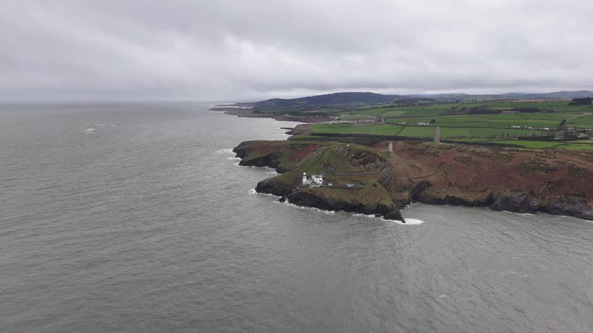 An aerial view captures the Wicklow Head Lighthouse perched on a rugged cliffside along the Irish coast on a dramatic, overcast day