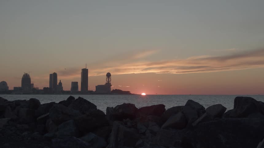 A tranquil sunset over the sea with a view of a distant city skyline from a rocky foreground