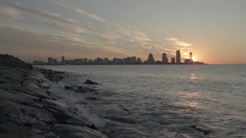 A slow tilt up from a rocky coast reveals a stunning city skyline across the ocean during a golden sunset