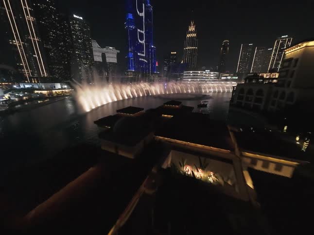 A nighttime aerial shot reveals the spectacular Dubai Fountain show in front of the iconic, illuminated Burj Khalifa