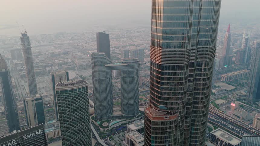 A slow aerial shot pulls back from the iconic Burj Khalifa skyscraper, revealing the expansive and hazy modern cityscape of Dubai at sunset