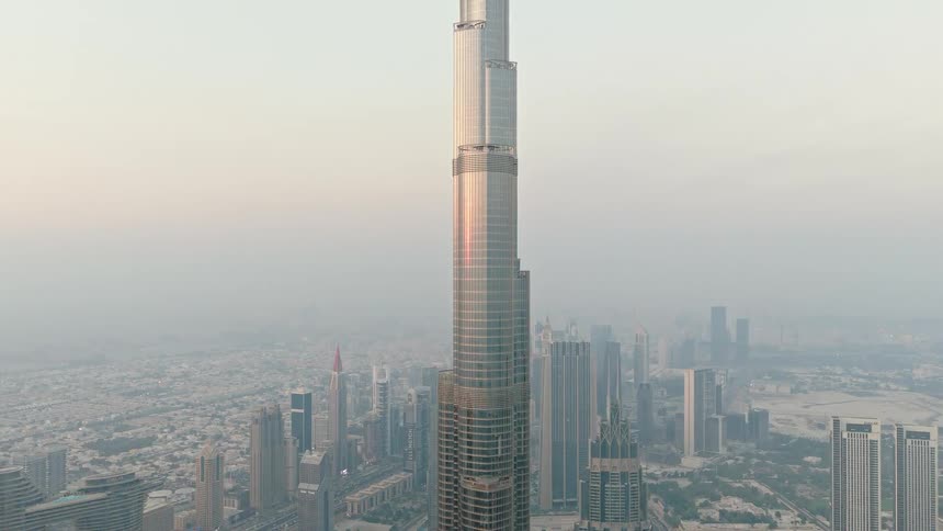 An aerial drone shot slowly ascends to reveal the full height of the iconic Burj Khalifa skyscraper against the vast, hazy Dubai skyline