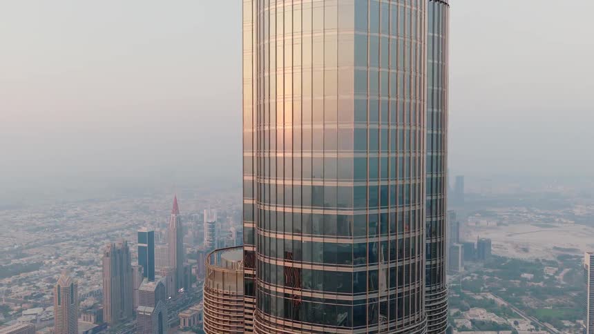 An aerial shot pulls back from the sun reflecting on the glass facade of the Burj Khalifa, revealing the vast, hazy cityscape of Dubai at golden hour