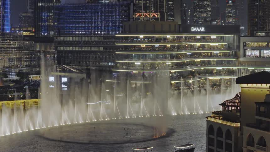 An enchanting aerial shot captures the dazzling Dubai Fountain show at night with boats on the water and the illuminated cityscape in the background