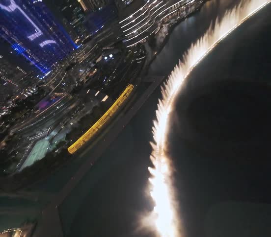 A dazzling nighttime aerial flight over the illuminated Dubai Fountain show, with the iconic city skyline as a backdrop