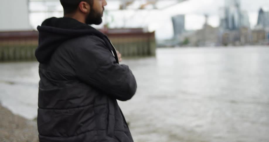 A young bearded man in a black jacket skips a stone across a river on a cloudy day in the city, filmed in slow motion