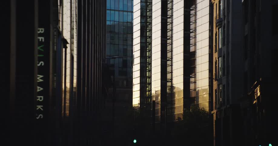 Slow pull-back shot reveals a shadowed London street where golden sunset light reflects off modern glass skyscrapers