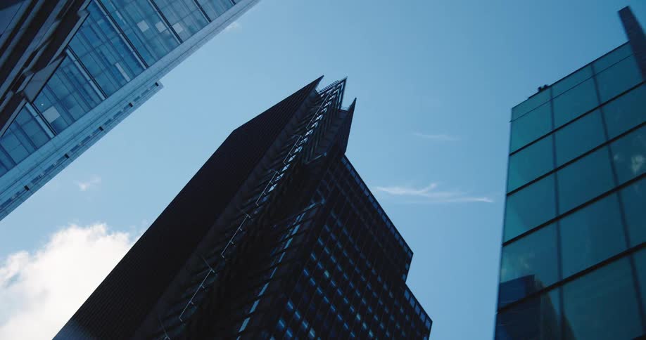 A low-angle pull-back shot reveals towering modern glass skyscrapers against a clear blue sky in a financial district