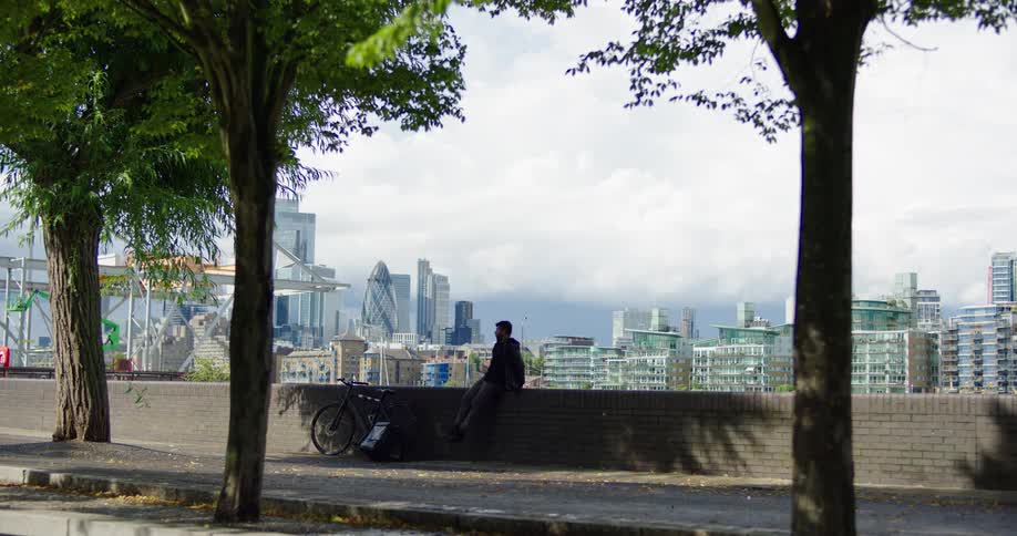 A food delivery cyclist takes a break on a brick wall, with a beautiful view of the London skyline in the background