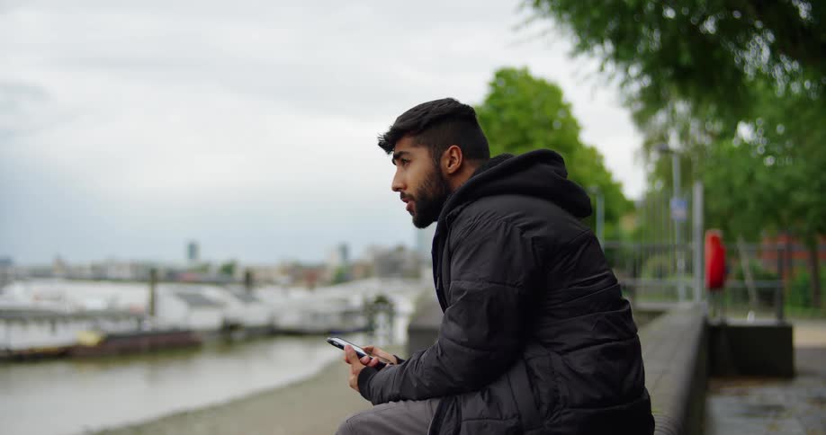 A young man in a black hooded jacket sits on a ledge by the river, thoughtfully looking at his smartphone on an overcast day