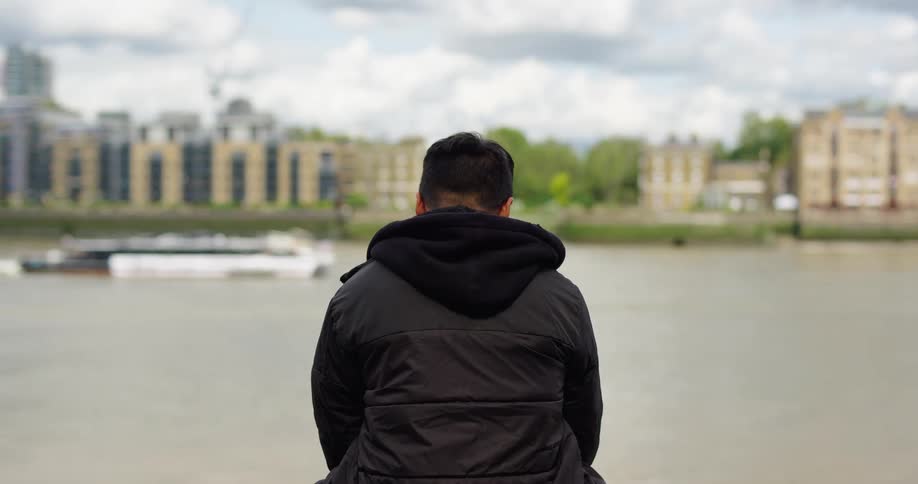 A man in a black hooded jacket sits with his back to the camera, looking out over a wide city river as boats pass by