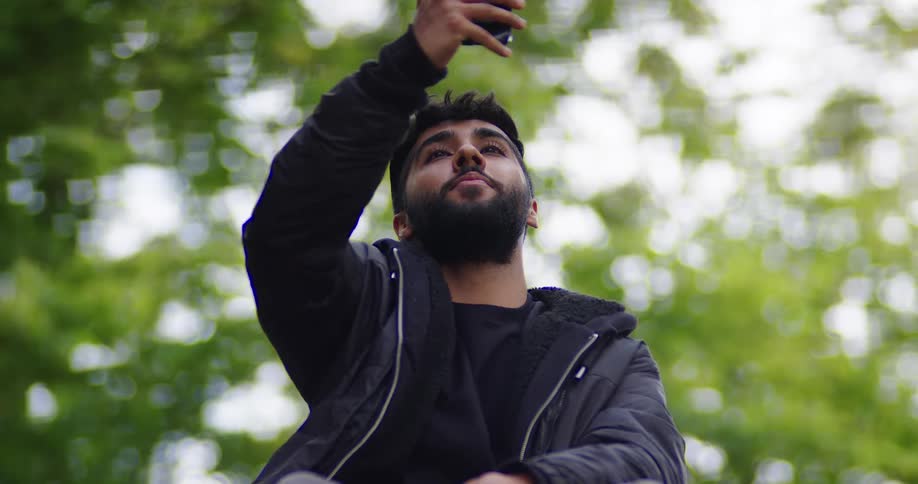 A young bearded man wearing a black jacket takes a selfie with his smartphone while sitting in a park
