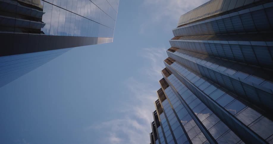 A worm's-eye view timelapse captures clouds moving across the blue sky reflected in two modern glass skyscrapers