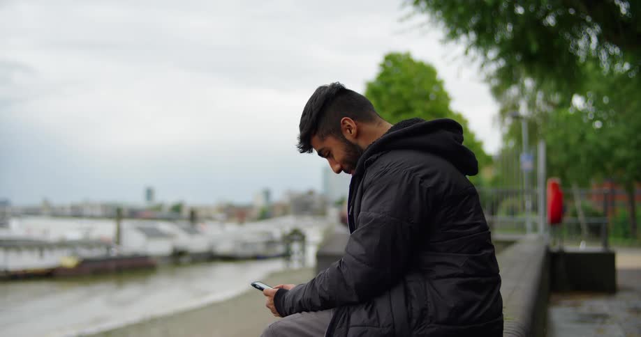 A young man enjoys a quiet moment using his phone while sitting by the water with a city skyline in the background