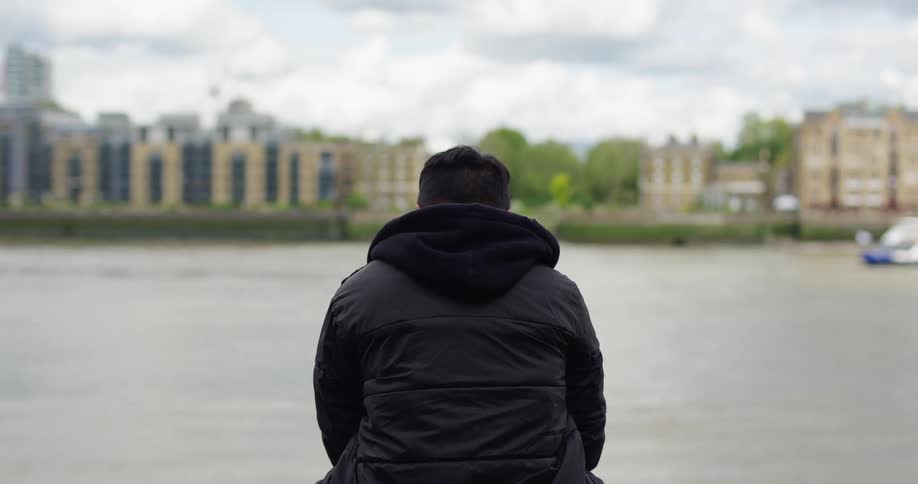 A lone figure in a black jacket sits pensively looking out over a city river on a cloudy day