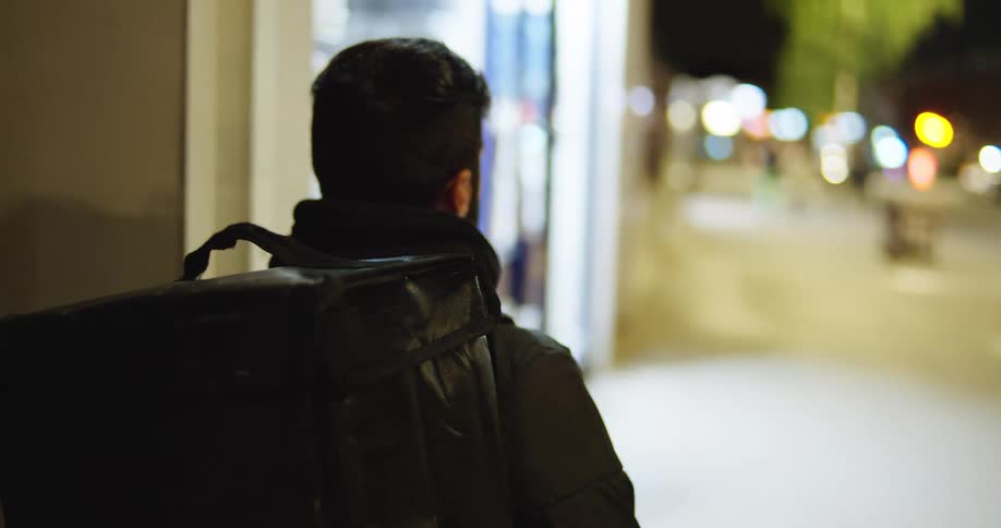 Male food delivery courier with an insulated backpack walks down a city street at night with bokeh lights