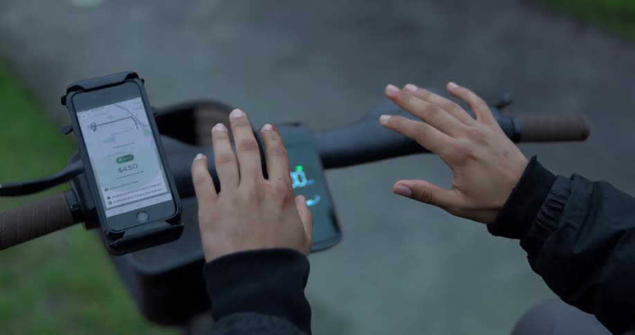 Close-up POV of a person with scraped hands over the handlebars of an electric scooter with a ride-sharing app on their phone