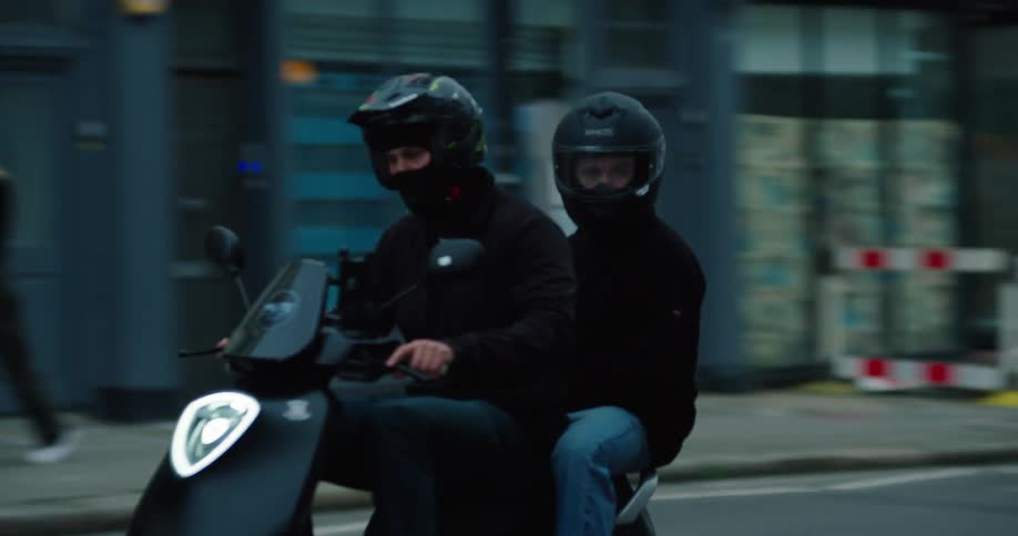 A slow-motion tracking shot of two men in helmets and face coverings riding a black scooter down a city street