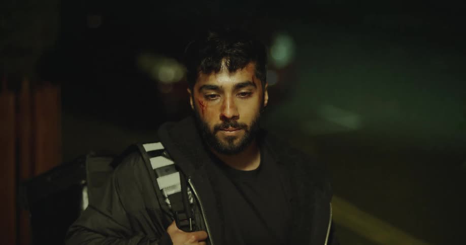 A bruised food delivery courier with cuts on his face looks sadly at the camera before walking away into the night