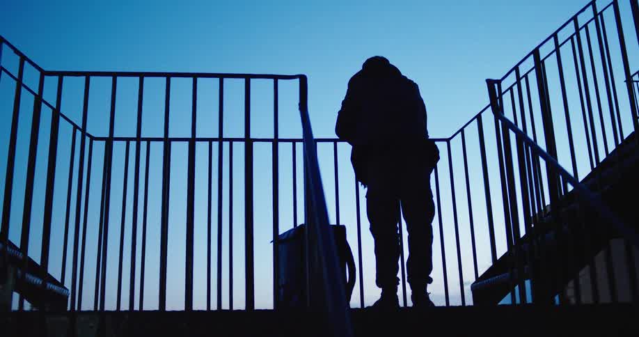 A silhouetted man with a backpack stands pensively on an outdoor metal staircase against a deep blue twilight sky