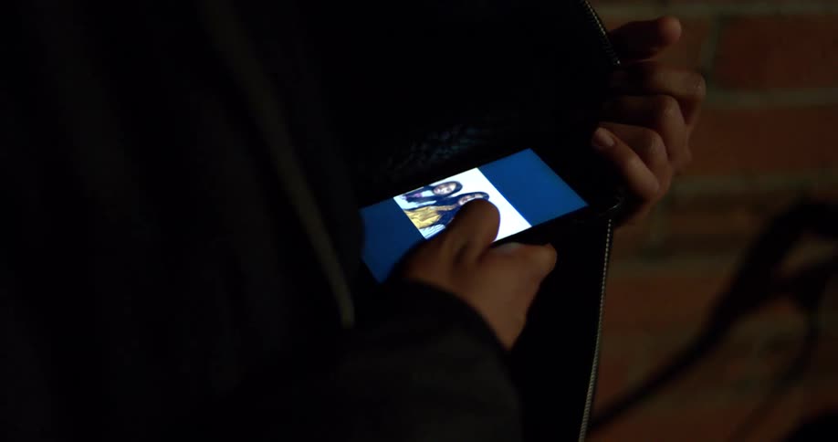 A person's hands secretly hold a smartphone inside a purse, the screen illuminating a photograph in the dark