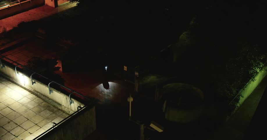 A high-angle time-lapse of a person holding an umbrella walking through a dark, rainy urban courtyard at night