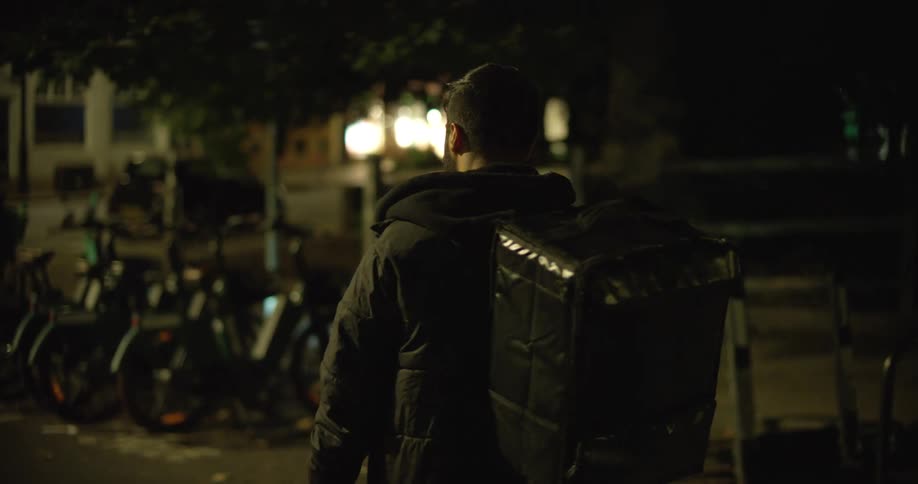 A food delivery courier wearing a thermal backpack walks through a city street at night to pick up a rental bicycle for his next job