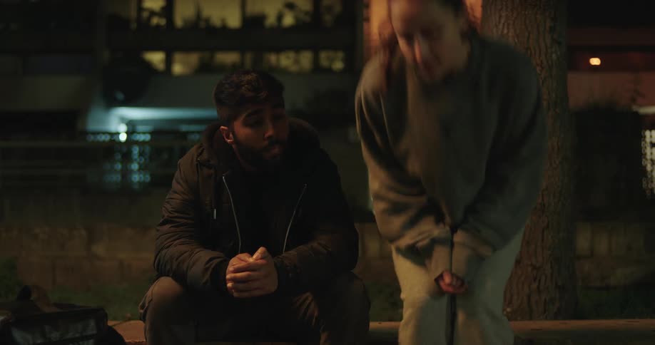 A young woman joins her somber friend sitting on a curb at night, and they sit together in quiet contemplation