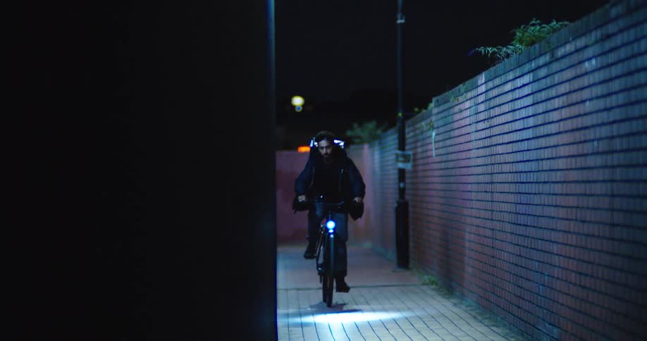 A man wearing a backpack rides his bicycle with a bright headlight through a dark city alleyway at night