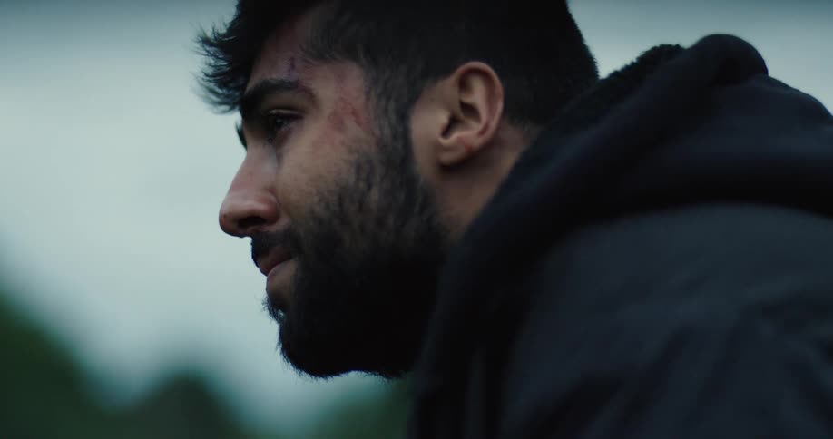 A close-up profile of an injured and emotional young man with a beard looking down as a single tear rolls down his cheek