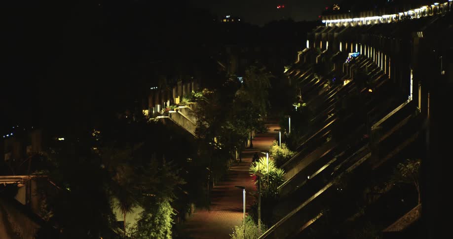 A lone cyclist rides down an illuminated path through a brutalist housing estate at night as the camera slowly zooms out