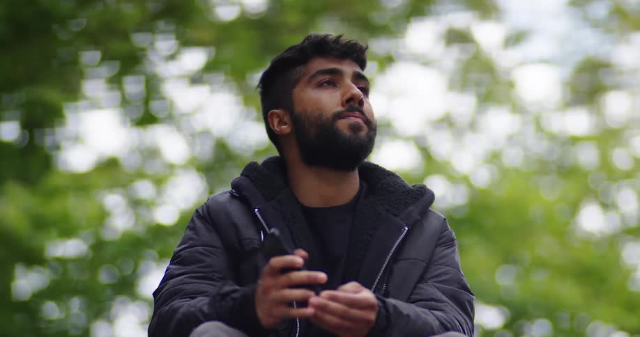 A pensive young man sends a voice message on his phone before taking a mindful break to appreciate the serene park scenery
