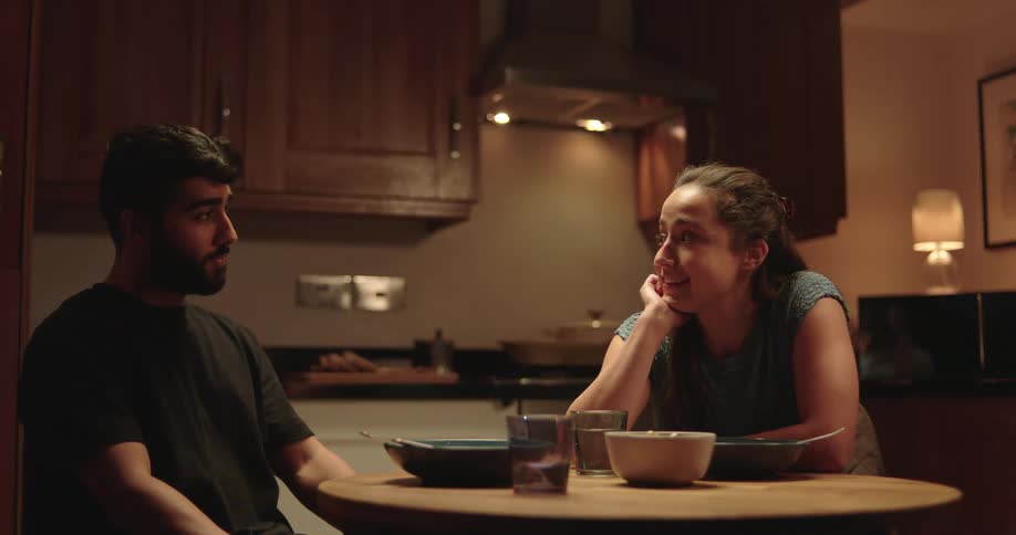 A young man confides in his girlfriend over dinner in their dimly lit kitchen