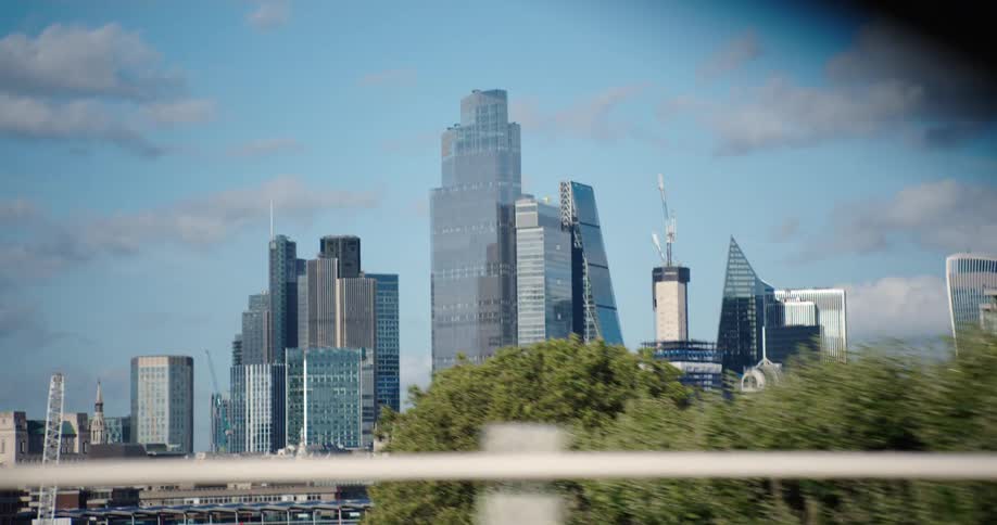 A tracking shot from a moving vehicle captures the modern skyscrapers of the London city skyline on a sunny day