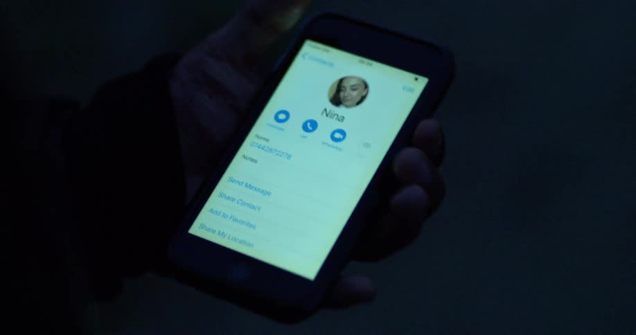 An injured hand holds a smartphone displaying a contact's information in a dark setting