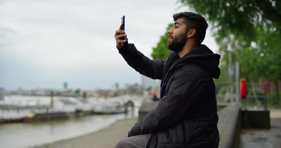A bearded young man in a black jacket sits by a city harbor on a cloudy day and takes a selfie with his smartphone