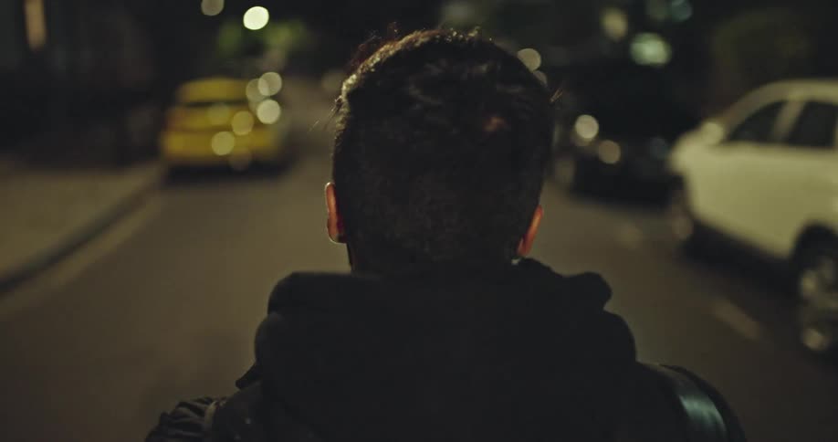 An over-the-shoulder tracking shot follows a man in a black hoodie as he walks down a dimly lit city street at night