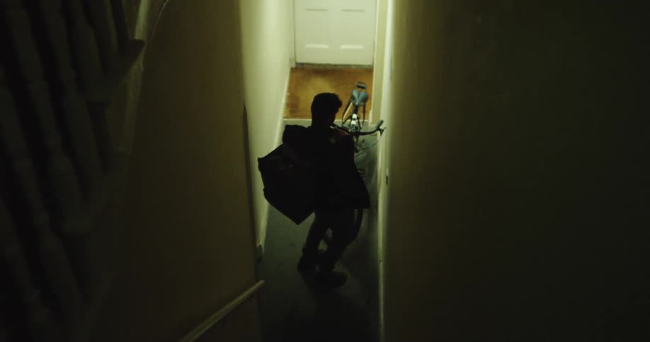 A food delivery cyclist grabs his bike and insulated bag from a dark apartment building hallway before heading out to work