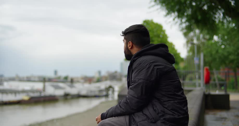 A young man eats a red lollipop and gets a burst of playful energy while sitting by a city harbor on an overcast day