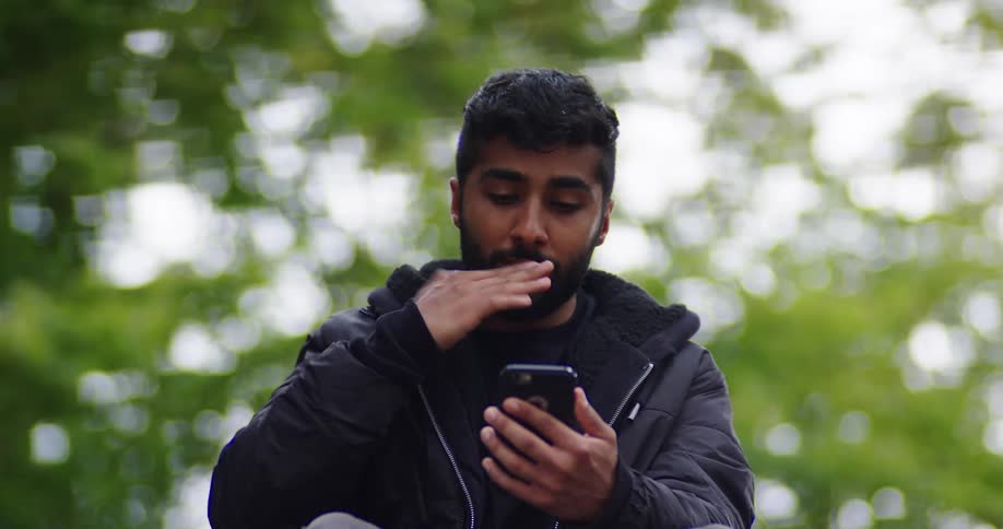 A young bearded man in a black jacket smiles while using his smartphone before looking up thoughtfully in a green park