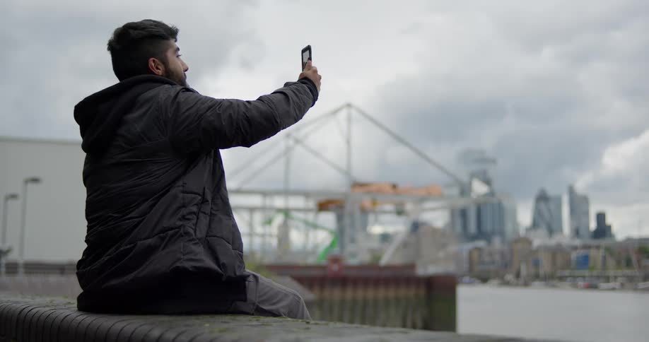 A man takes a selfie on his smartphone while sitting by a river, with the London city skyline visible in the background